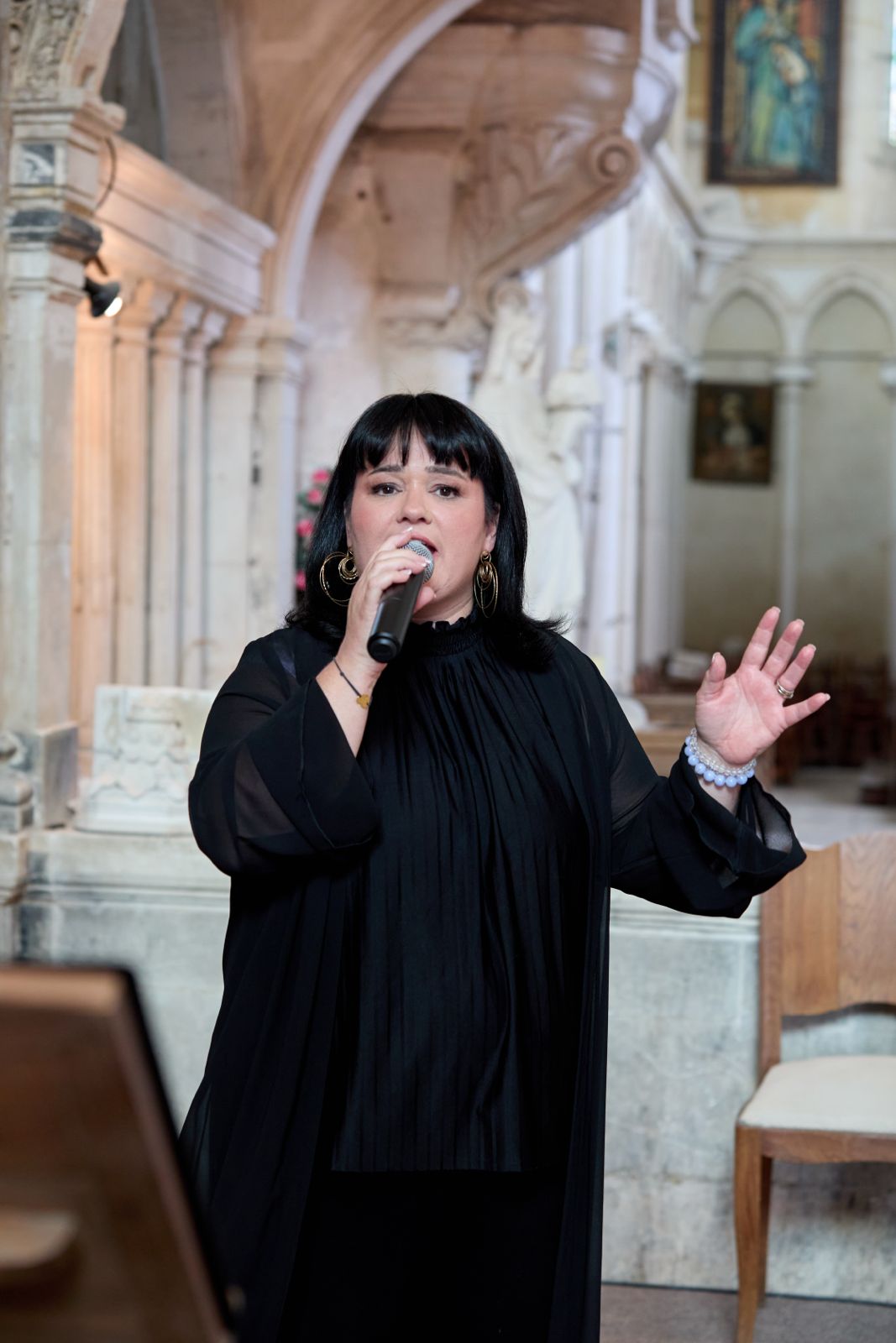 Image d'Ingrid chantant face à la caméra avec le micro à la bouche. Elle fait un signe de la main. Elle se trouve dans une église.